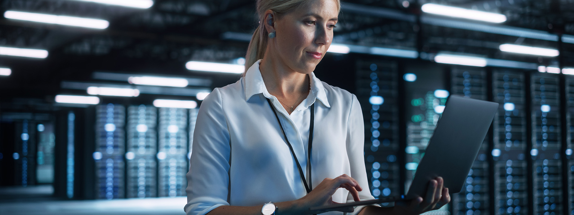 Eine Frau in weißem Hemd hält einen Laptop in einem Serverraum.