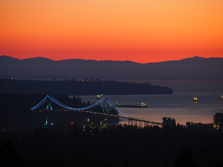 Die Lions Gate Bridge während des Sonnenuntergangs, im Hintergrund ist der berühmte Stanley Park und eine Gebirgskette auf Vancouver Island zu sehen
