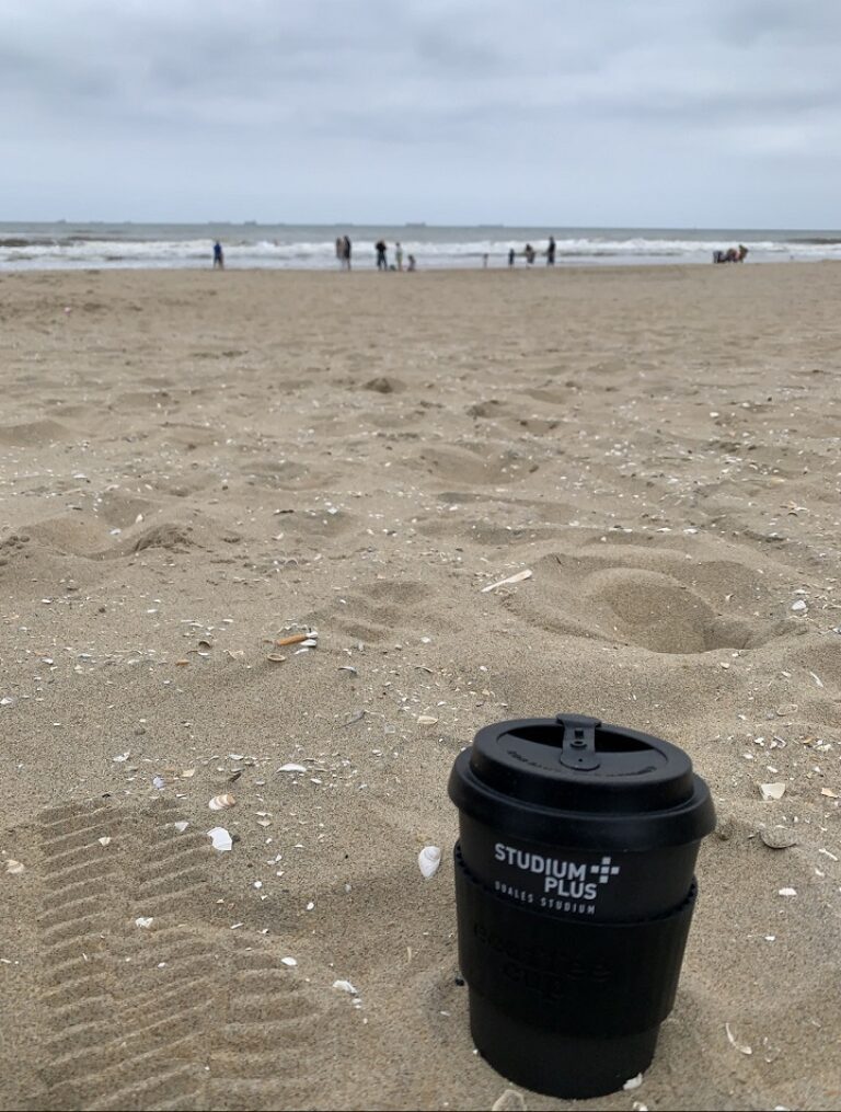 Eine Kaffeetasse am Strand mit Menschen im Hintergrund.