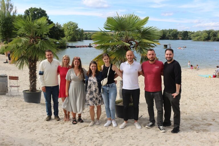 Eine Gruppe junger Menschen in sommerlicher Kleidung steht am Strand neben einem See.