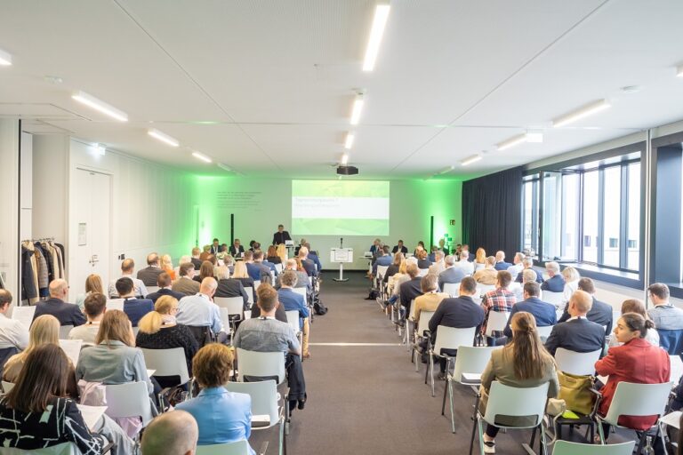 Mitglieder sitzen in großem Saal auf Stühlen mit Blickrichtung auf die Redner und die Präsentation im vorderen Bereich.