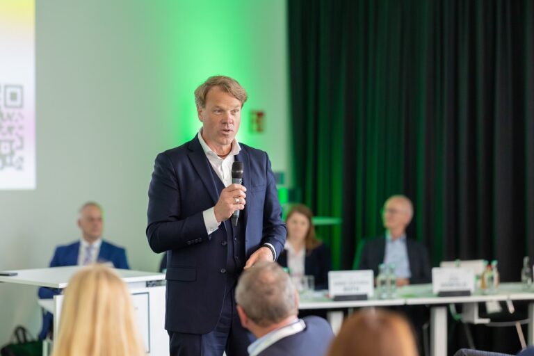 Ein Professor in einem Anzug steht vor den Mitgliedern der Konferenz und spricht engagiert in ein Mikrofon.